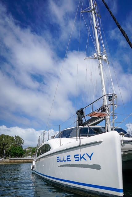 A white catamaran named "BLUE SKY" is docked in calm water under a partly cloudy blue sky, with trees and a shoreline visible in the background.