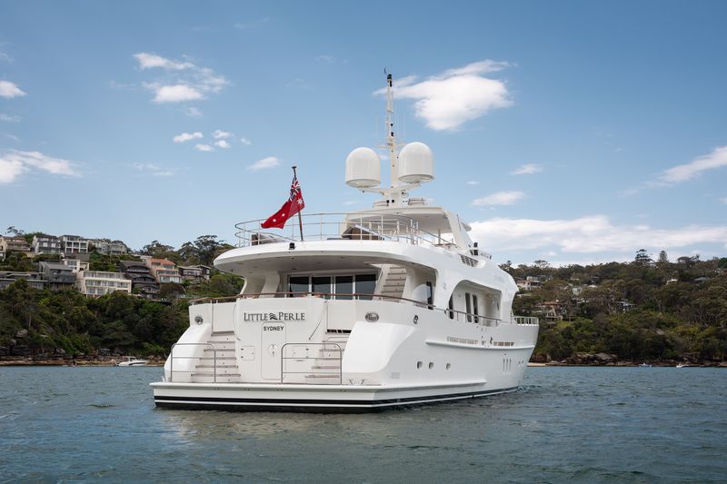 A large white yacht named "Little Pearle" with a red flag is anchored on calm water near a hilly coastline with houses and trees under a partly cloudy sky.
