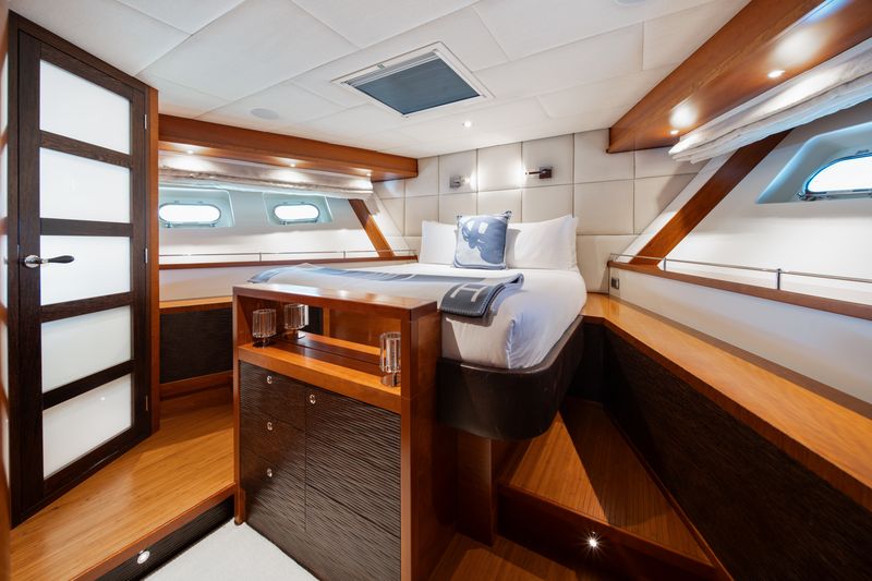 A luxurious yacht bedroom with a large bed, white bedding, wood-paneled walls and furniture, two small windows, and modern lighting, creating a bright and elegant atmosphere.