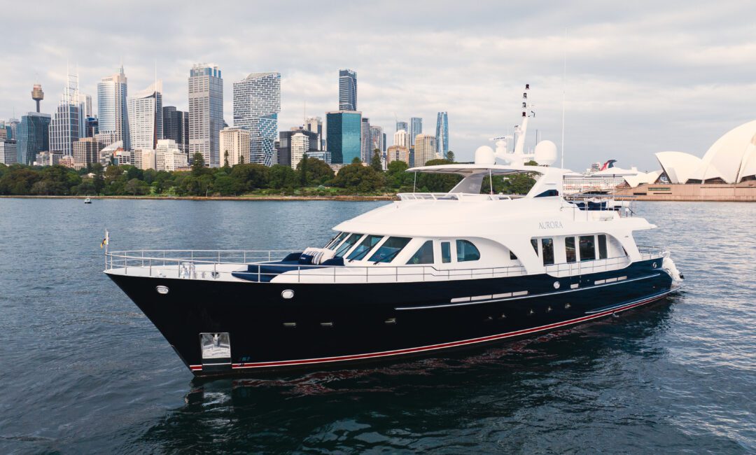 A large luxury yacht sails on the water with a modern city skyline and the Sydney Opera House in the background under a cloudy sky.