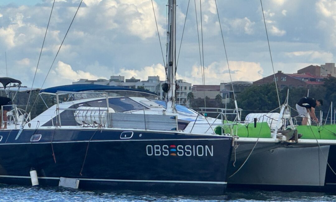 A blue sailboat named "OBSESSION" docked on the water, with buildings and trees visible in the background under a partly cloudy sky.