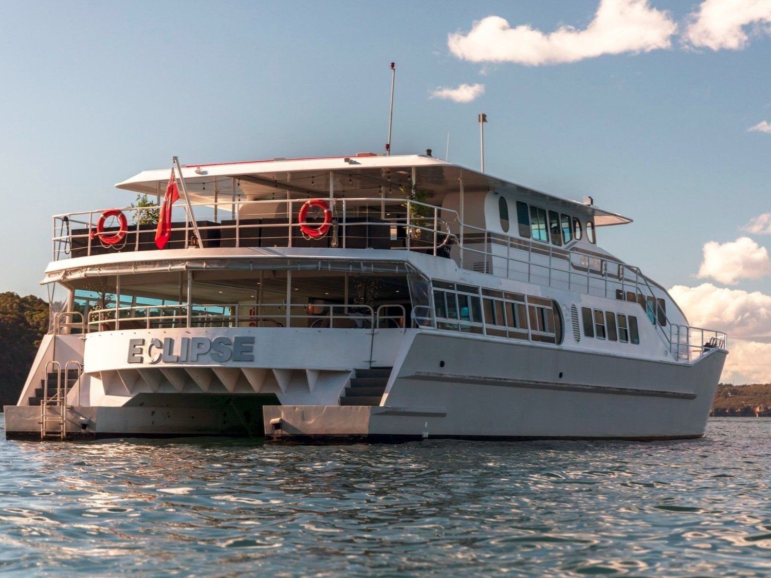 A large white yacht named "ECLIPSE" is anchored on calm water under a partly cloudy sky. The vessel has multiple decks, railings with lifebuoys, and a red flag at the stern.