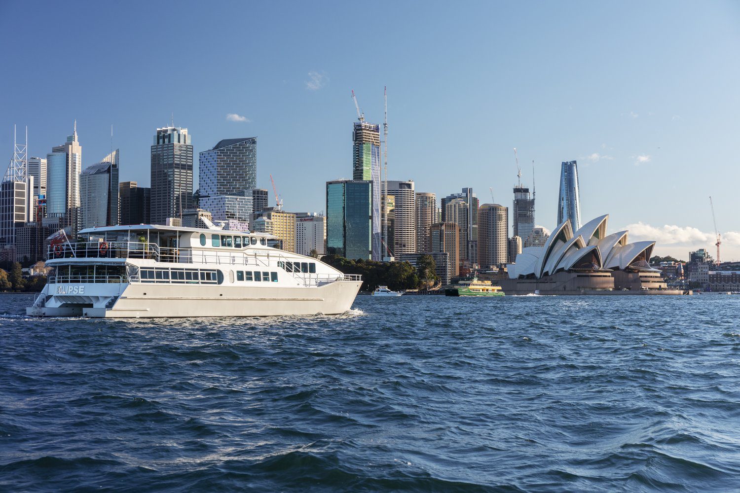 A white boat sails on blue water with Sydney's city skyline and the iconic Sydney Opera House visible in the background under a clear sky.