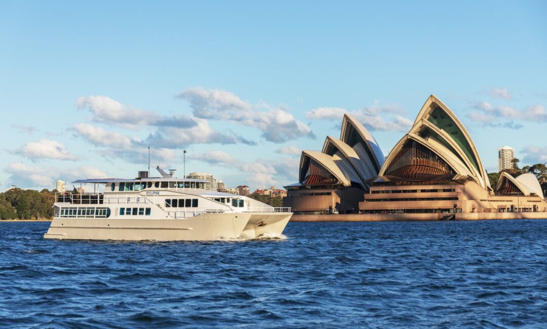 A white boat sails on blue water near the Sydney Opera House in Australia, with clear skies and scattered clouds in the background.