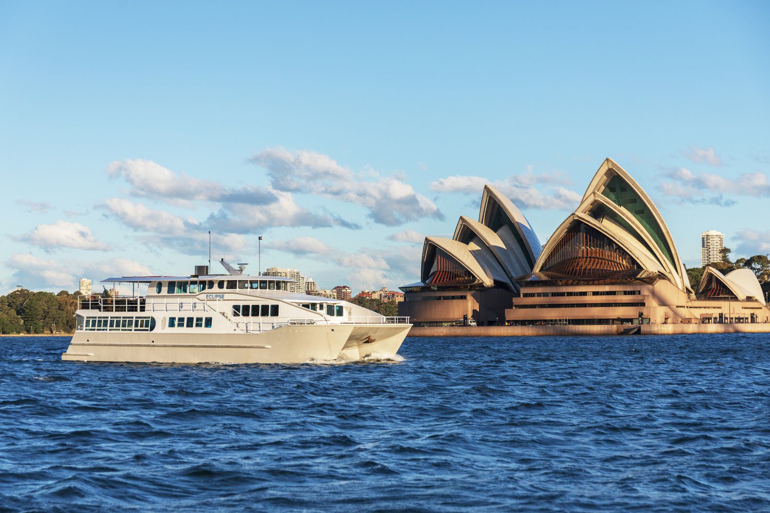 A white boat sails on blue water near the Sydney Opera House in Australia, with clear skies and scattered clouds in the background.