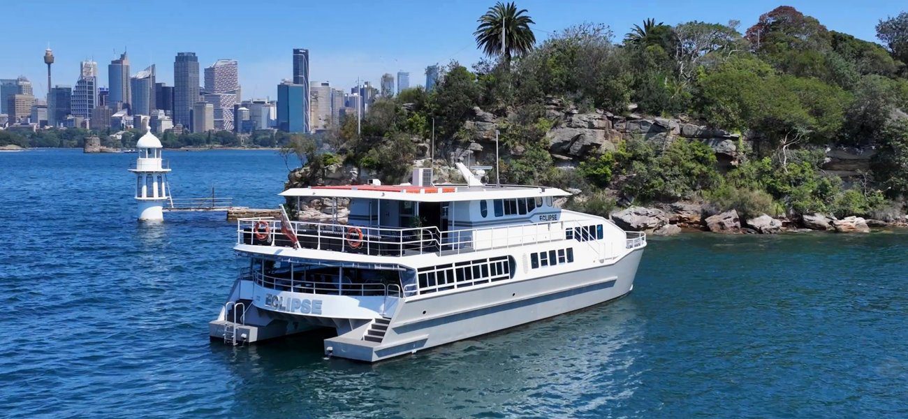 A white catamaran named Eclipse is anchored near a rocky, tree-covered island, with a city skyline and blue water in the background under a clear sky.