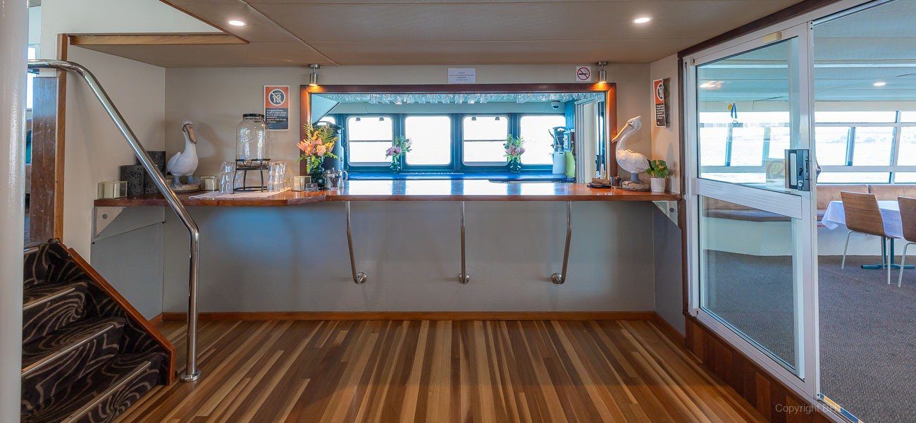 A modern boat interior with wood-striped flooring, a counter with flowers and coffee setup, stairs on the left, and large windows overlooking water in the background.