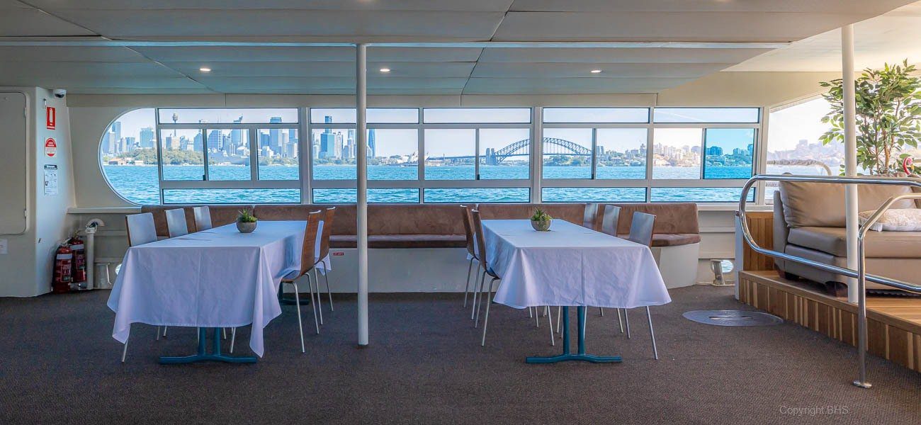 Interior of a boat with tables and chairs covered in white cloths, large windows offering a clear view of Sydney’s skyline and the Sydney Harbour Bridge across the water.