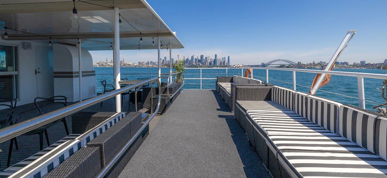 A view from the deck of a boat with striped cushioned seating, looking out toward a city skyline and a bridge over blue water under a clear sky.