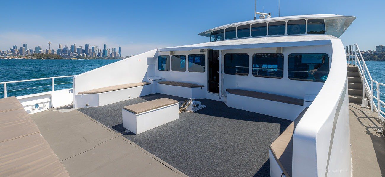 Spacious upper deck of a modern boat with cushioned seating, benches, and a table, overlooking a city skyline across the water under a clear blue sky.