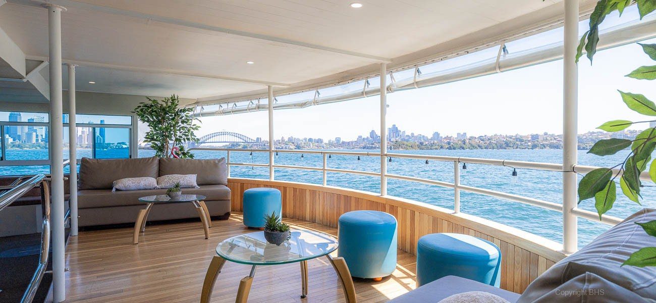 A spacious boat lounge with modern seating, plants, and small tables overlooks the water. Large windows reveal a city skyline and a landmark bridge under a clear sky.