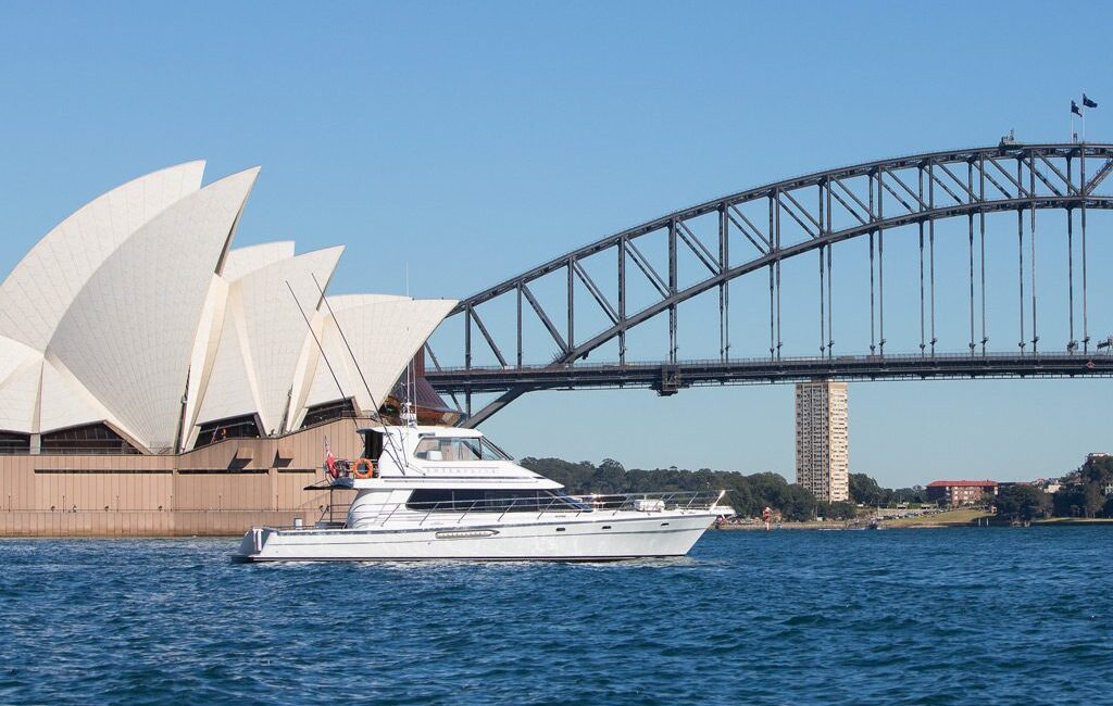 A white yacht sails on blue water in front of the Sydney Opera House, with the Sydney Harbour Bridge in the background, under a clear blue sky.