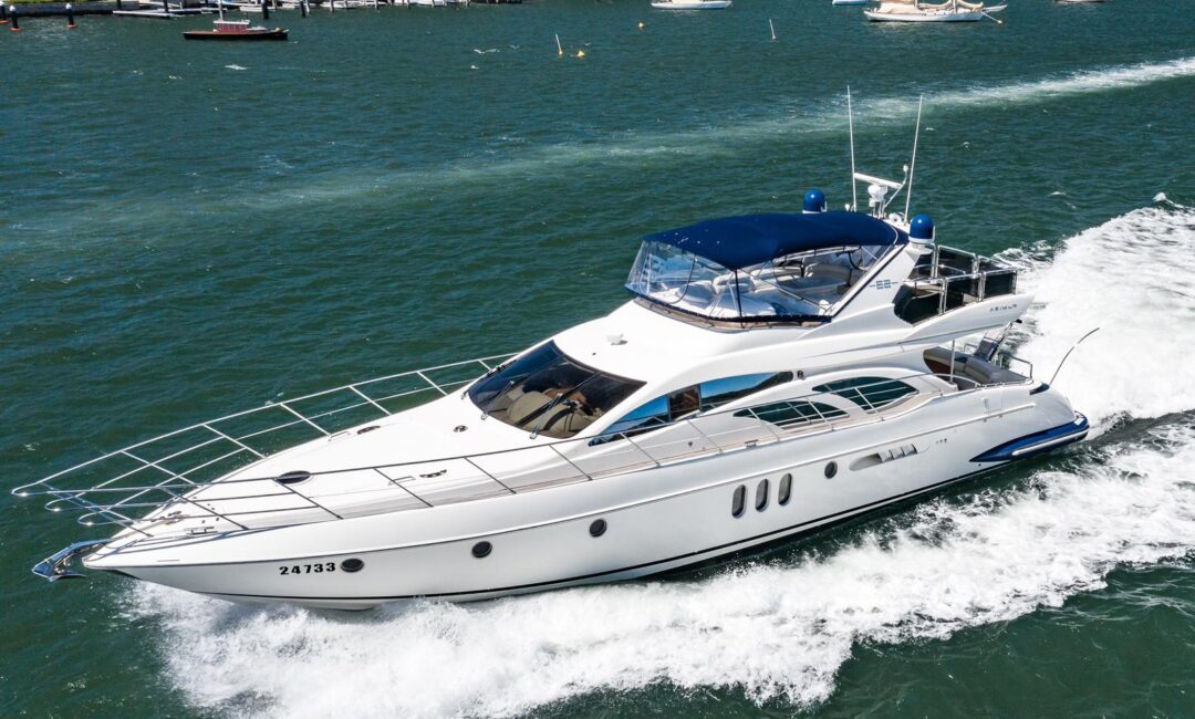 A sleek white yacht cruises through a green-blue harbor, creating a trail of white waves, with houses, trees, and several anchored boats visible along the shoreline in the background.