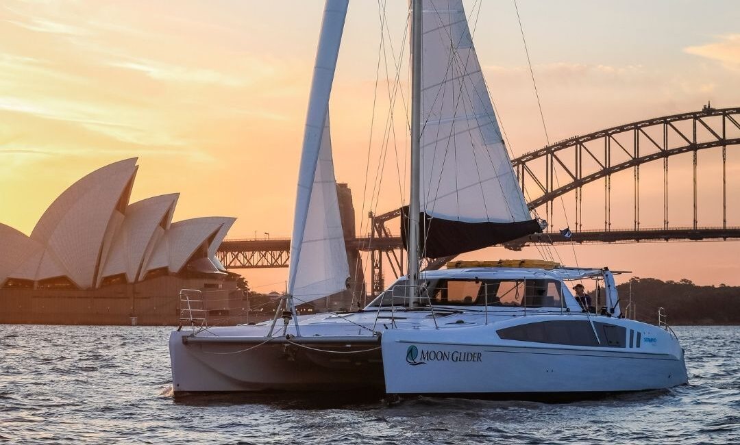 A white sailing catamaran named "Moon Glider" floats on the water at sunset with the Sydney Opera House and Sydney Harbour Bridge visible in the background.