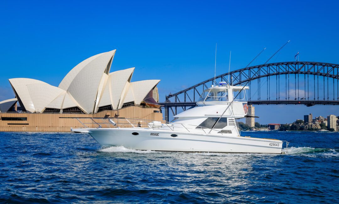 A white motor yacht cruises on Sydney Harbour with the Sydney Opera House and Harbour Bridge in the background under a clear blue sky.