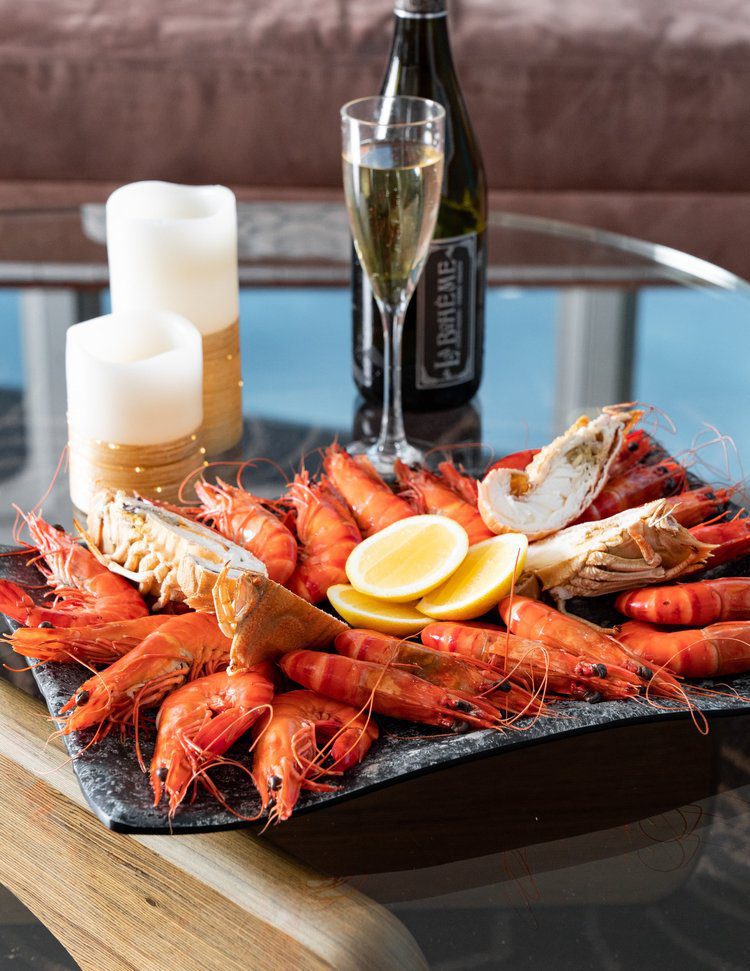 A platter of cooked prawns and lobster with lemon wedges sits on a glass table next to a glass and bottle of champagne and three lit candles.