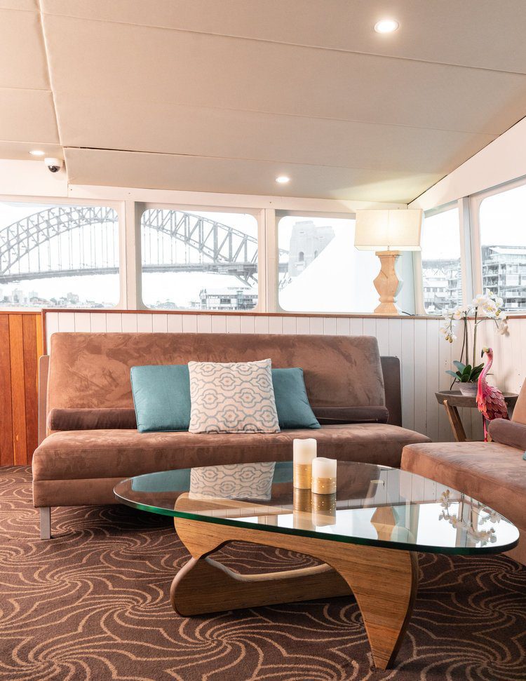 A cozy living area with brown sofas, blue and patterned cushions, a glass coffee table, candles, a lamp, and a view of the Sydney Harbour Bridge through large windows.