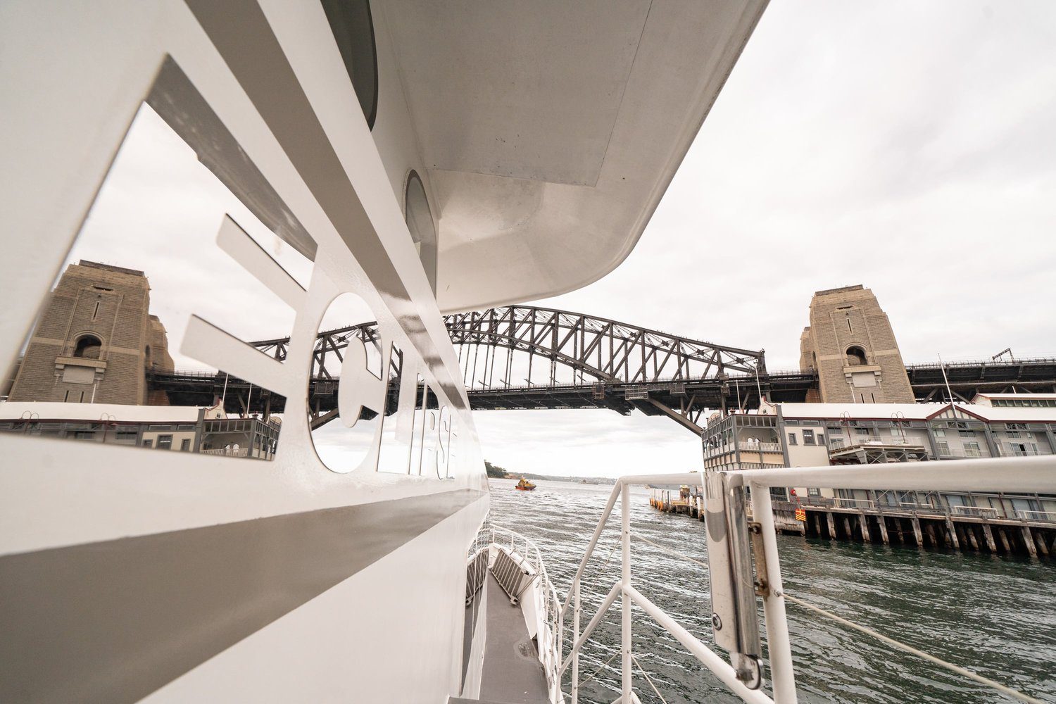 A view of the Sydney Harbour Bridge from the deck of a boat, with the bridge's stone pylons and waterfront buildings visible under a cloudy sky.