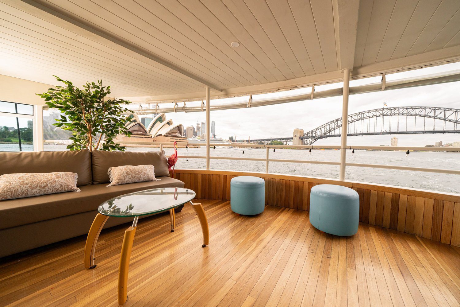 A modern boat interior with a brown couch, glass coffee table, and two blue ottomans, overlooking Sydney Opera House and Sydney Harbour Bridge through large windows.