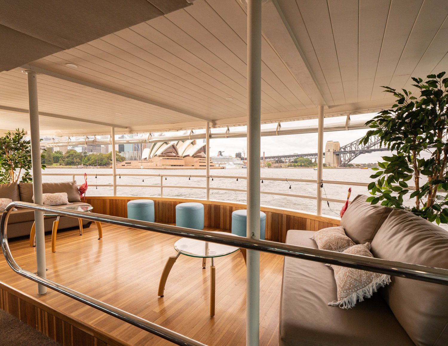 Modern boat deck with cushioned seating, glass tables, and potted plants overlooking the water. Sydney Opera House and Harbour Bridge are visible in the background through open railings.