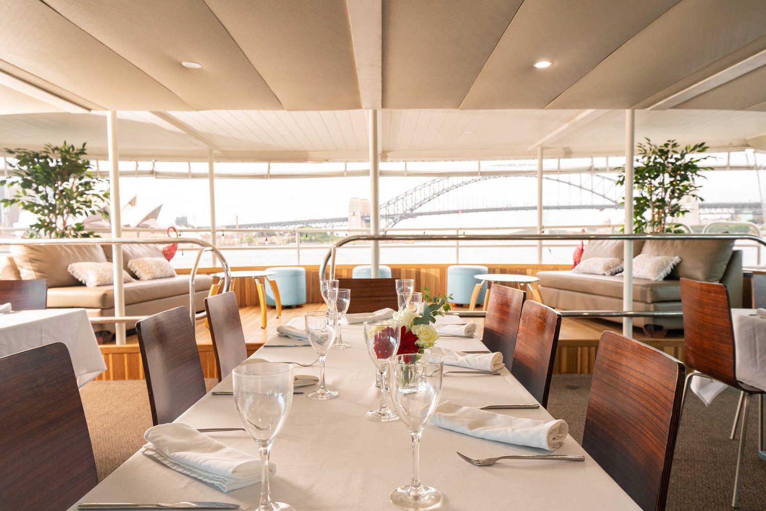 A dining area on a boat with set tables, elegant glassware, and white napkins, overlooking Sydney Harbour, with the Sydney Opera House and Harbour Bridge visible through large windows.