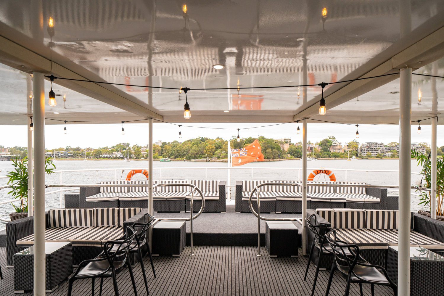 Covered outdoor deck on a boat with striped seating, black chairs, and string lights. A red flag is flying at the stern, with water and a tree-lined shoreline visible in the background.