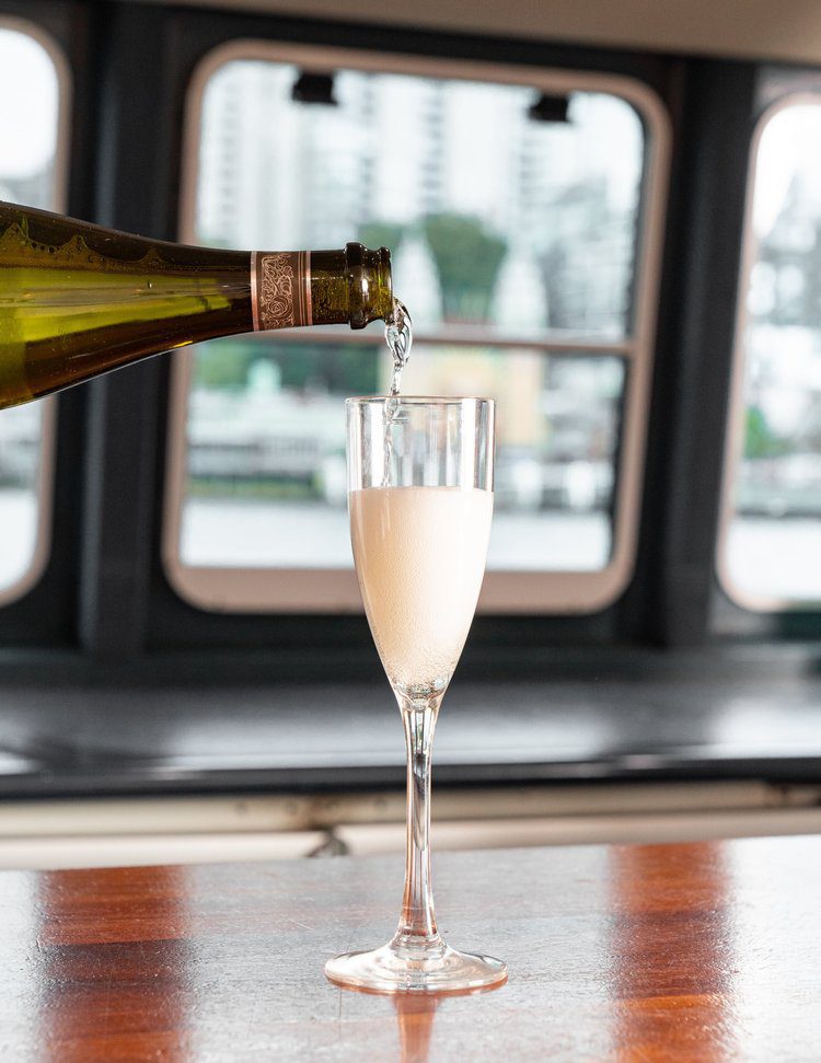 A bottle pours sparkling wine into a tall champagne flute on a wooden table, with blurred windows and a cityscape visible in the background.