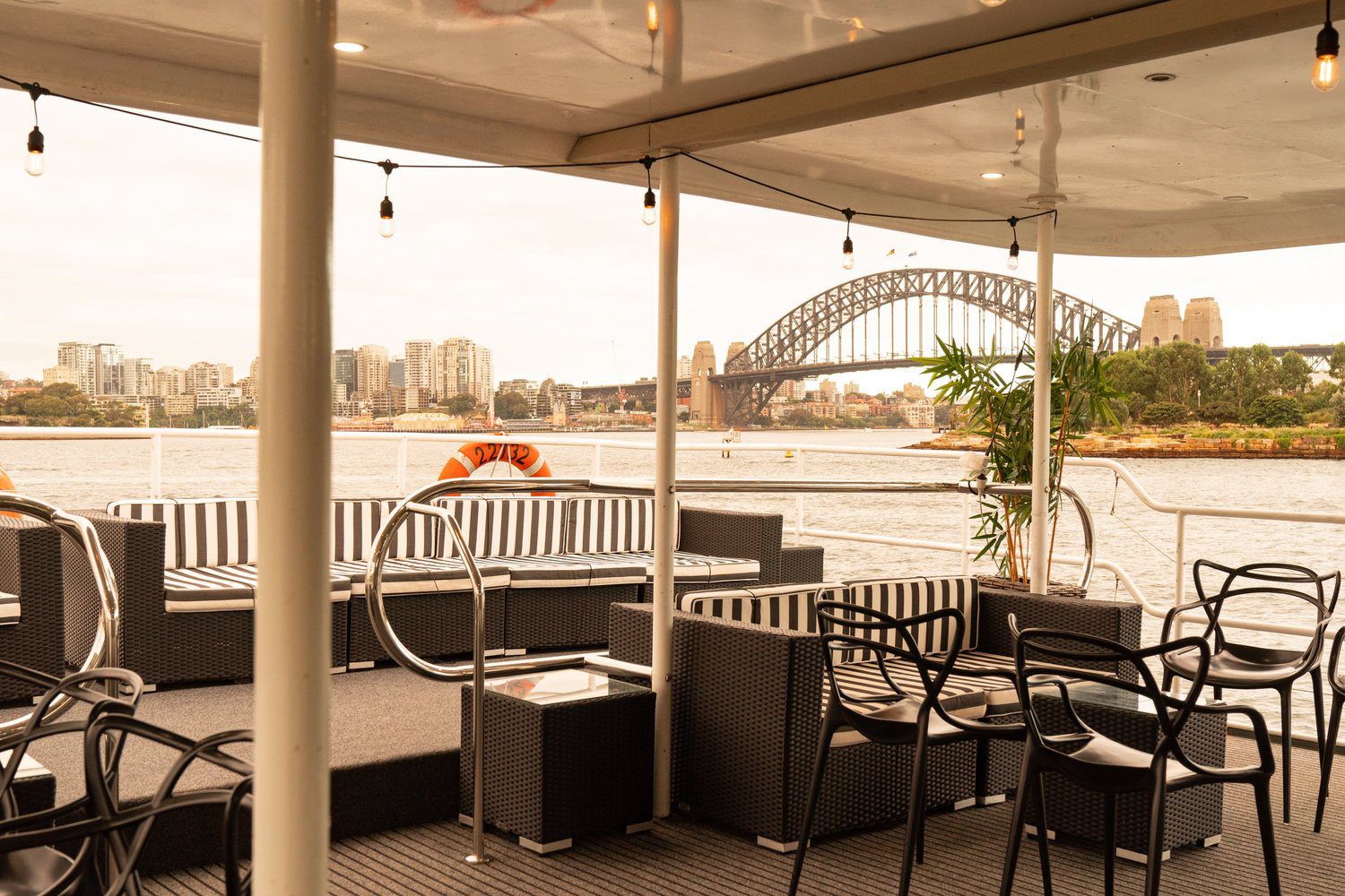 Outdoor seating area on a boat with black and white furniture, overlooking the Sydney Harbour Bridge and city skyline on a cloudy day.
