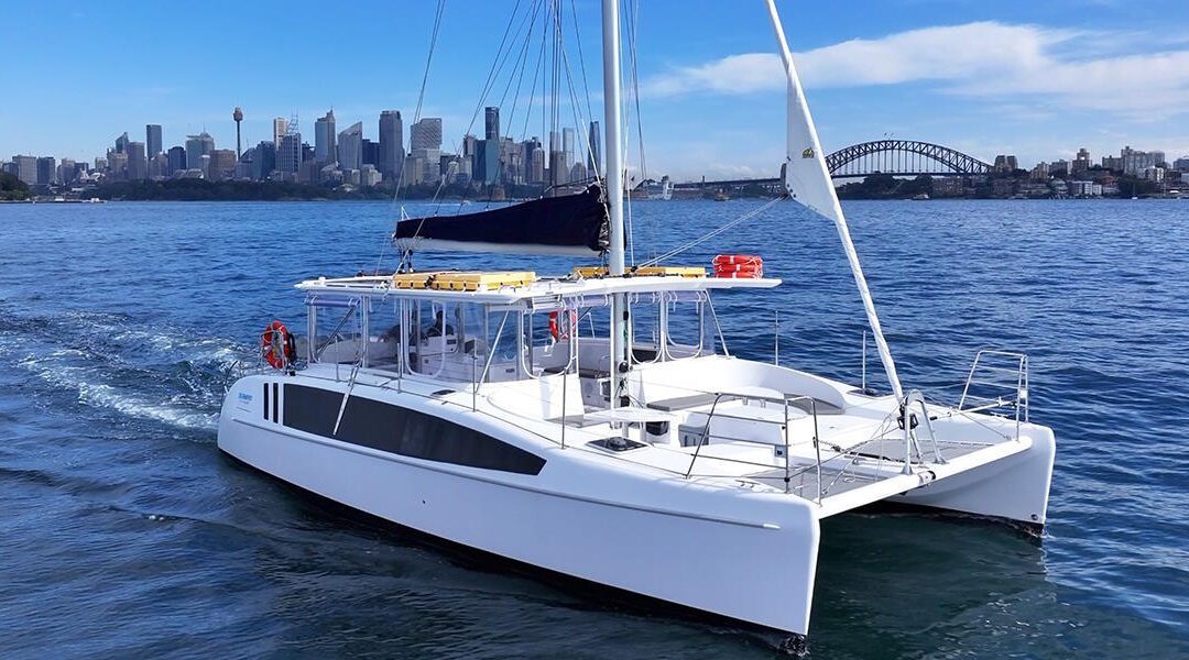 A white catamaran sails on blue water with a city skyline and a large bridge visible in the background under a partly cloudy sky.