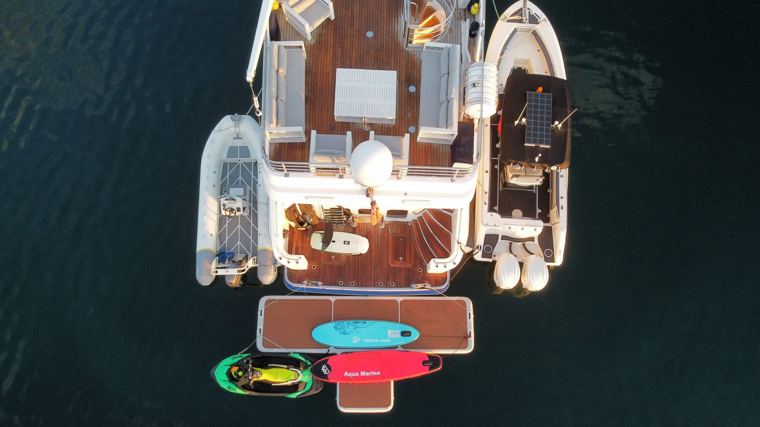 Aerial view of a yacht docked beside a smaller boat, with two paddleboards—one blue and one pink—and a kayak floating near the back deck on calm water.