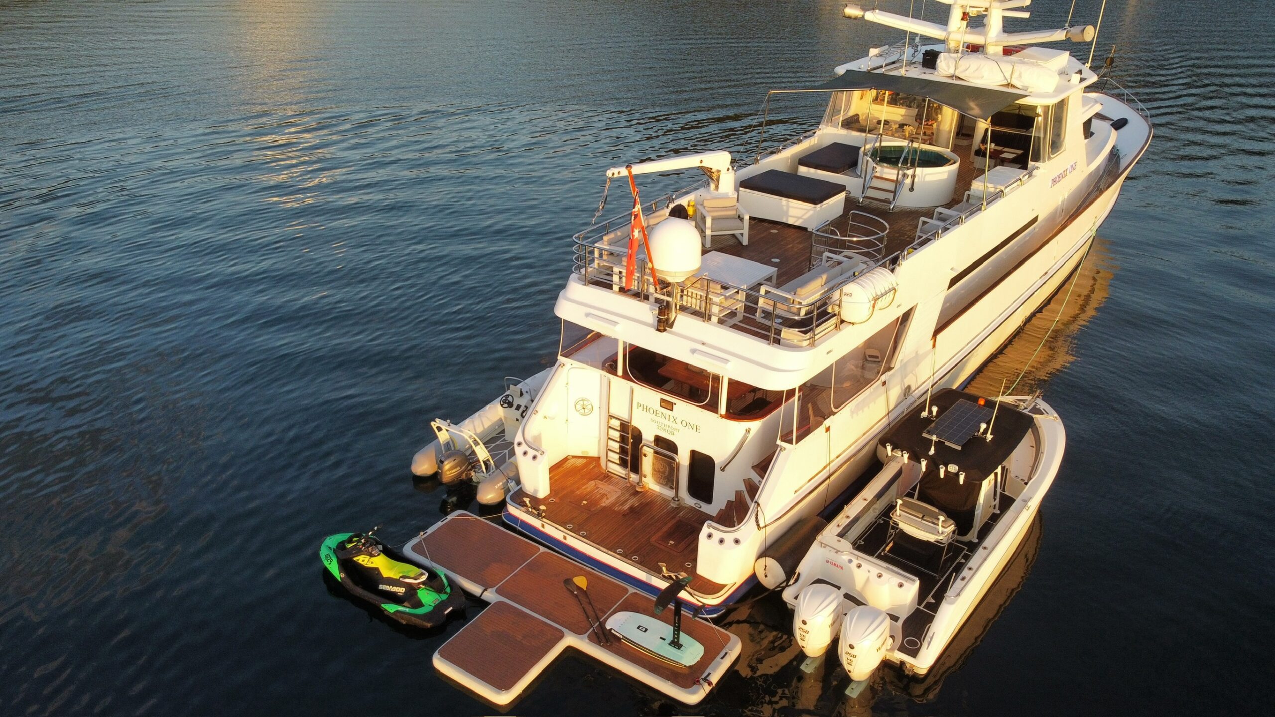 A large white yacht is docked on calm water, with its rear deck featuring a small jet ski, floating platforms, and an inflatable dinghy tied alongside. The sun casts a warm light on the scene.