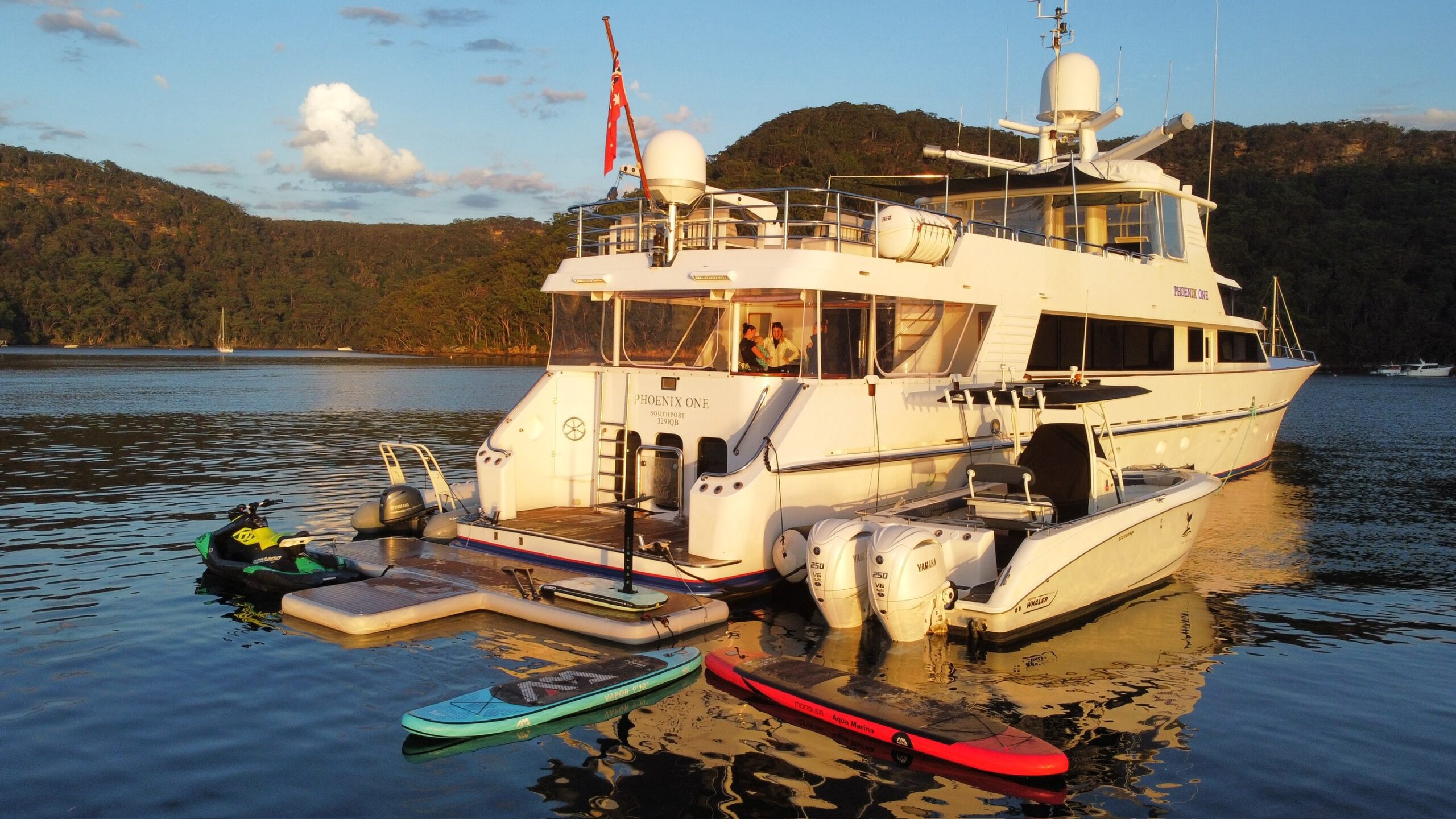 A large white yacht anchored in calm water near forested hills, with a smaller boat, paddleboards, and jet skis beside it, under a partly cloudy sky at sunset.