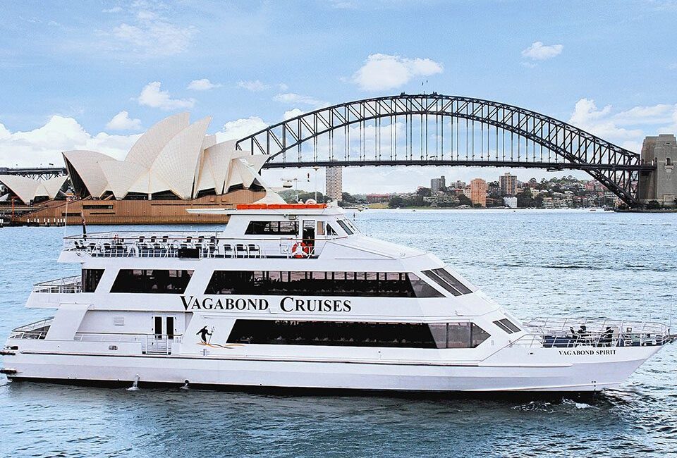 A white Vagabond Cruises boat sails in Sydney Harbour, with the Sydney Opera House and the Sydney Harbour Bridge visible in the background under a partly cloudy sky.