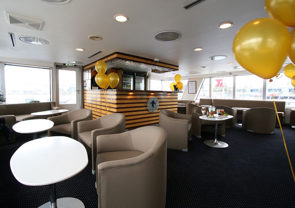 A modern lounge area with beige armchairs, white tables, and yellow balloons, featuring a wooden bar at the center and large windows bringing in natural light. Drinks and snacks are set on a few tables.