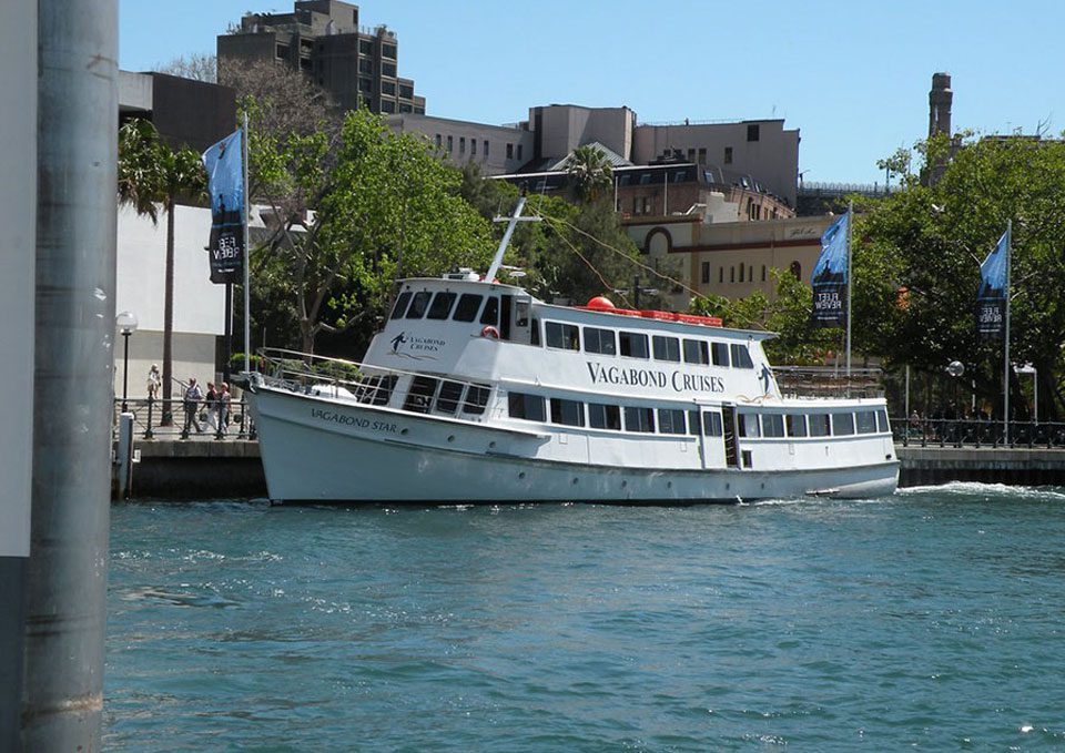 A white cruise boat named "Vagabond Star" from Vagabond Cruises is docked by a pier next to a tree-lined waterfront with buildings in the background and blue water in the foreground.