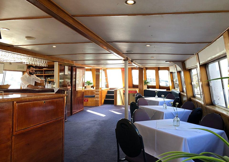 Interior of a boat dining area with wooden paneling, tables covered in white cloths, dark chairs, small flower vases, large windows, and a bartender standing behind a wooden counter. Natural light fills the space.