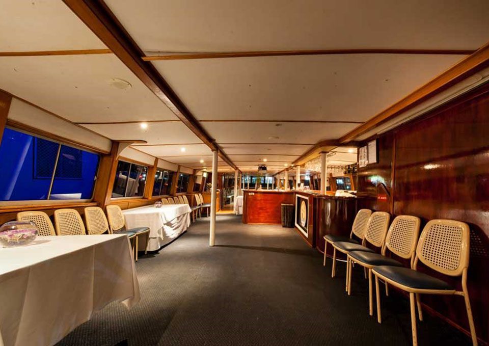 Interior of a boat or yacht with empty chairs lined up along the wood-paneled walls, large windows on one side, and a table with a white tablecloth on the left. The space is warmly lit and appears ready for an event.
