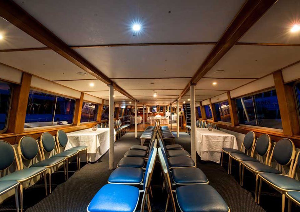 Rows of blue cushioned chairs are arranged facing each other on a boat's lower deck, with large windows along the sides and white tablecloth-covered tables near the back, under warm ceiling lights.