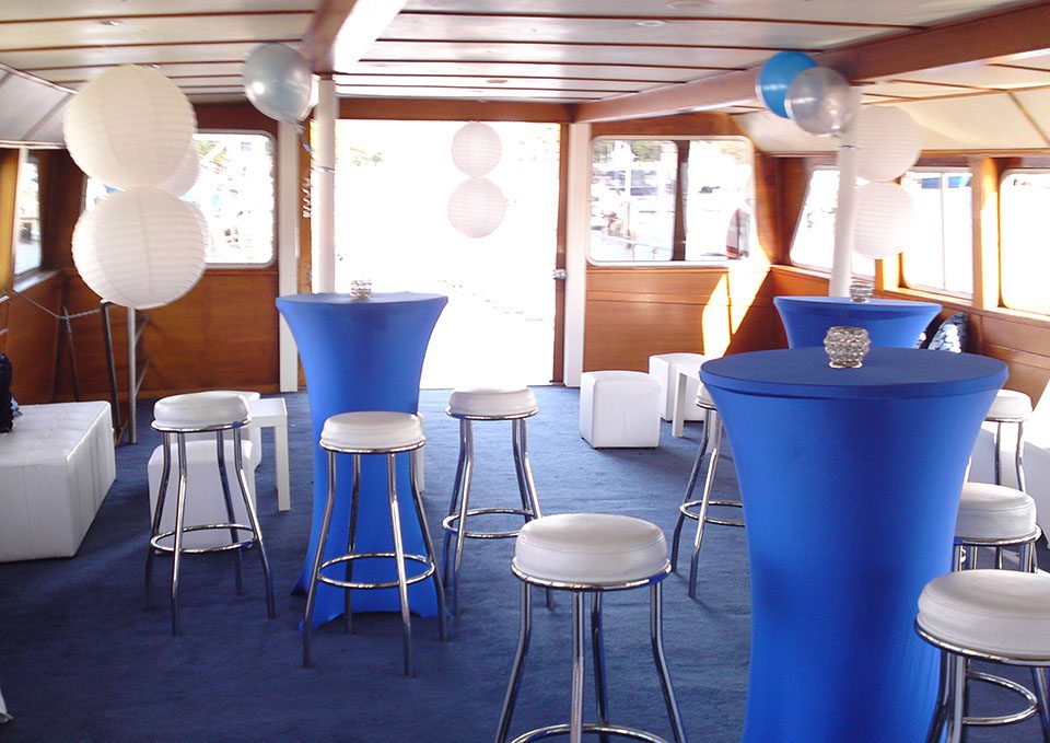 A boat interior decorated for an event with blue tablecloths on tall cocktail tables, white barstools, white cube seats, and white paper lanterns and balloons, all set on a blue carpet. Sunlight streams through large windows.
