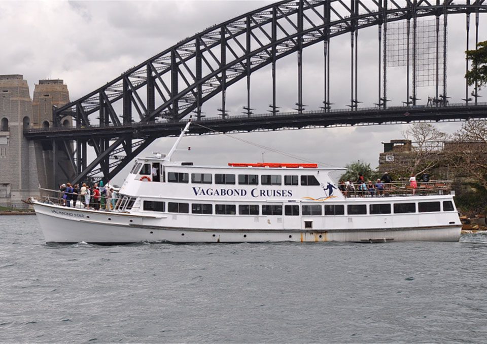 A white sightseeing boat labeled "Vagabond Cruises" sails on the water with passengers on deck; the Sydney Harbour Bridge and cloudy sky are visible in the background.