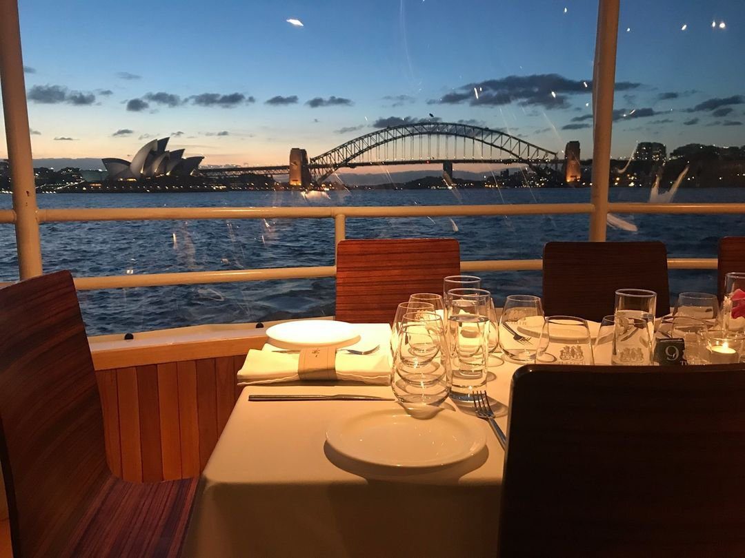 A dining table set with plates and glasses on a boat, with a view of Sydney Harbour, featuring the Sydney Opera House and Harbour Bridge at sunset in the background.