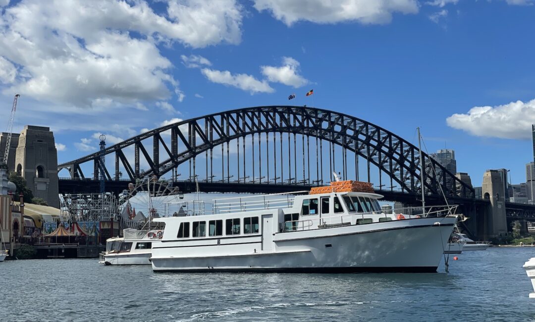 A white passenger boat floats on the water with the Sydney Harbour Bridge in the background under a blue sky with scattered clouds. Other boats and city structures are also visible.