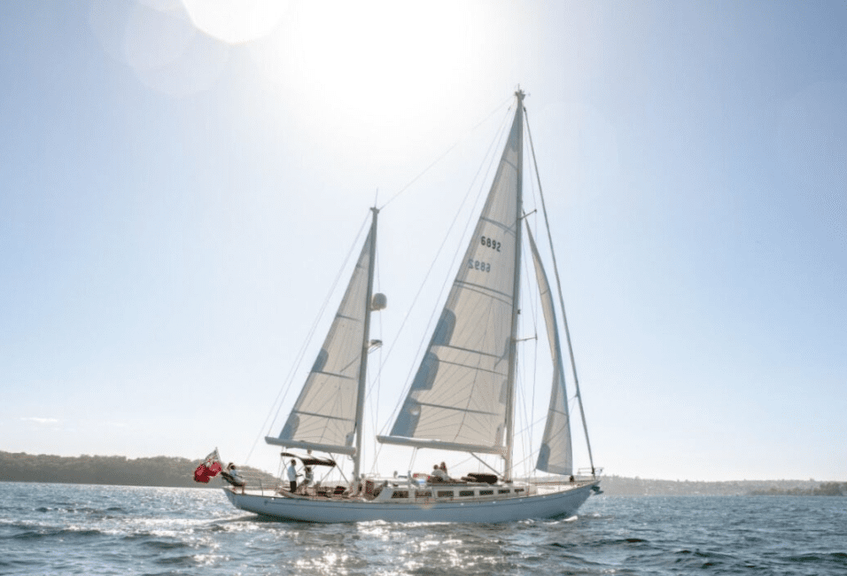 The Sir Thomas Sopwith, a white sailboat with two sails and a red flag at the stern, glides across a calm blue sea under a clear sky, with several people on board and the coastline visible in the background.