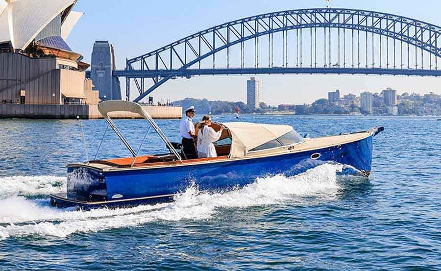 A small blue motorboat with three people cruises on the water near the Sydney Opera House and Sydney Harbour Bridge on a sunny day.