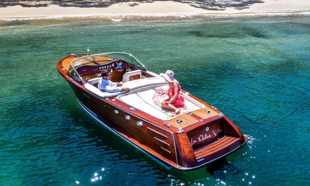 A sleek wooden motorboat with two people onboard, a man at the wheel and a woman in a pink dress and hat, floats on clear turquoise water near a sandy shore lined with trees.