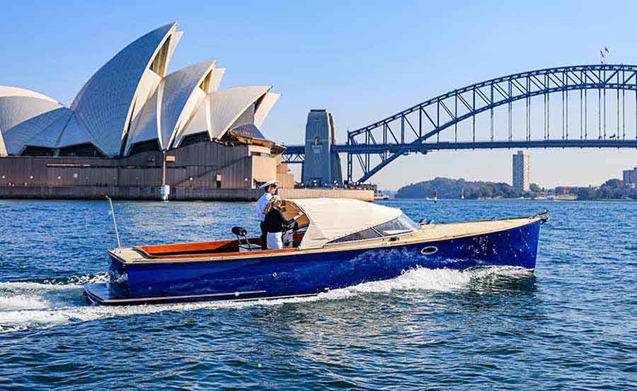 A small blue motorboat cruises on the water in front of the Sydney Opera House and Sydney Harbour Bridge under a clear blue sky.