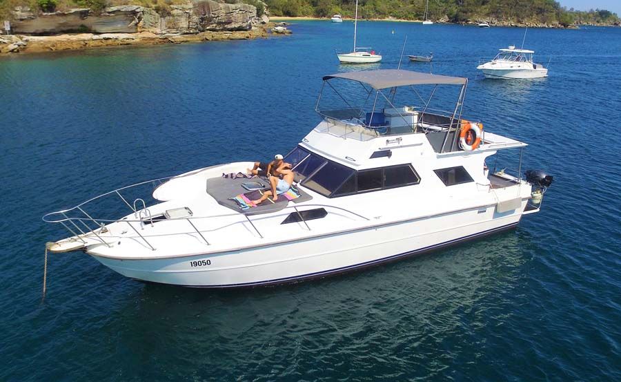The Mayfair, a white motor yacht, floats on calm blue water near the shore. Two people sunbathe on the bow as other boats are anchored nearby against rocky, tree-lined land.