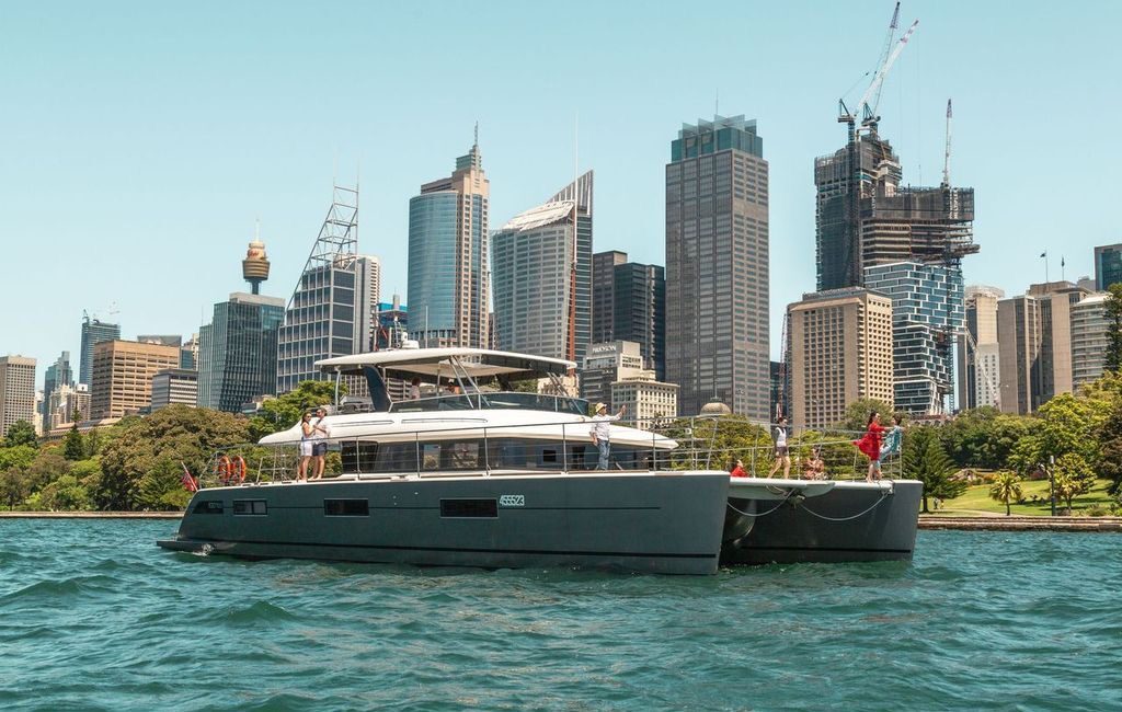 A modern catamaran yacht sails on blue water with a city skyline and high-rise buildings in the background on a clear, sunny day.