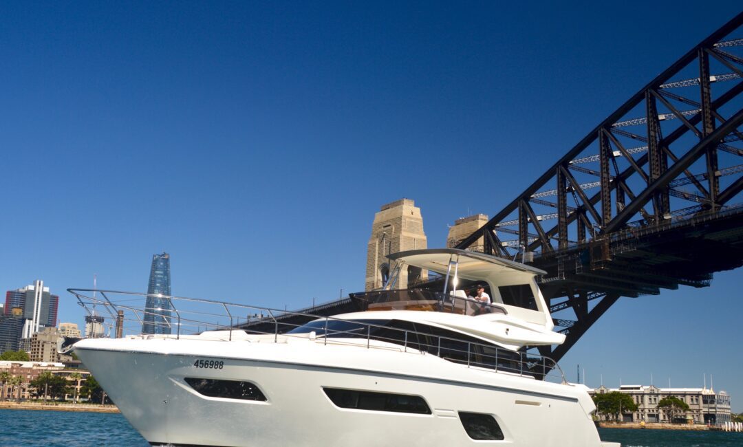 A white yacht sails near the Sydney Harbour Bridge on a sunny day, with modern city buildings visible in the background against a clear blue sky.