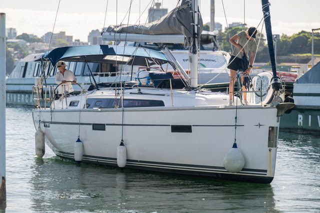 A white sailboat with two people aboard is docked in a marina, with other boats and city buildings visible in the background. One person is at the helm while the other works near the mast.